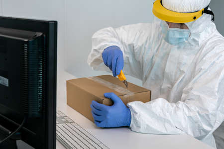 Doctor in protective suit and plastic face shield opens a box of face masks during Covid19. Man...