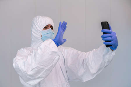 Male nurse say hello with the hand to patient talking on the mobile phone in interior of clinic setup amid Coronavirus pandemic. Telemedicine concept with smartphone in hospital during covid-19.の写真素材