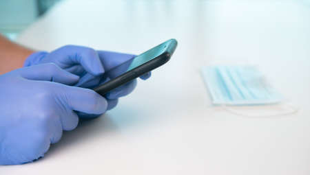 A person with gloves is typing on smartphone on a white table. Nearby is a surgical mask. Concept of...