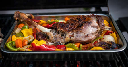 Chef cooking a delicious leg of lamb on a barbecue with colors peppers and potatoes in a metal tray....