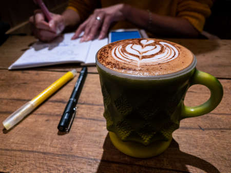 View of woman drinking cappuccino cup with heart shape latte art foam at coffeehouse. Student sitting at wooden table and writing lecture in notebook. Enjoy studying indoor cafe shop and taking notes.の写真素材