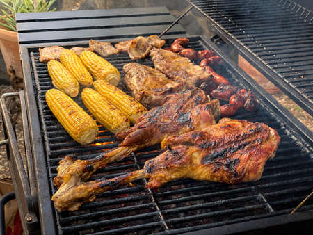 Cooking delicious leg of lamb and pork ribs with corn on the cob in a metal tray grilled. Cook a sausage of chorizo meat and tasty corns on BBQ grill. Chef cooking pork in backyard of a houseの写真素材