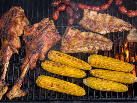 Cooking delicious leg of lamb and pork ribs with corn on the cob in a metal tray grilled. Cook a sausage of chorizo meat and tasty corns on BBQ grill. Chef cooking pork in backyard of a houseの写真素材