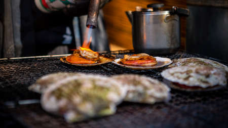 Vendor cooking scallops on steel grill in asian fish market street at Japan. People cooked popular seafood grilling BBQ. Flame fire of charcoal. Famous marketplace Tokyo. Delicious japanese foodの写真素材