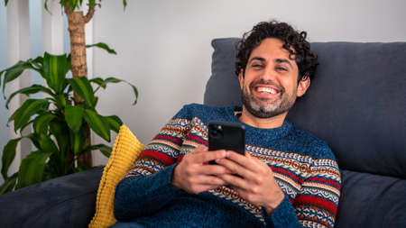Portrait of happy caucasian man smiling while using smartphone and sitting on couch at beautiful home interior. Caucasian man relaxing with phone device on sofa of house.の写真素材