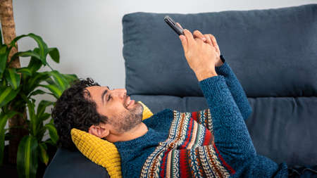 Portrait of happy caucasian man smiling while using smartphone and lying down on couch at beautiful home interior. Caucasian man relaxing with phone device on sofa of house.の写真素材