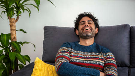 Young handsome man with beard wearing winter sweater with happy face smiling looking at the camera and sitting on sofa of home. Positive person.の写真素材