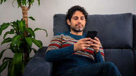 Cheerful handsome man using his smart phone while sitting on the couch at home. Confident bearded man is typing on smartphone at sofa of house.の写真素材