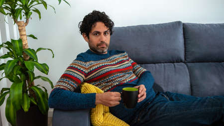 Caucasian man with beard drinking morning expresso coffee and looking at the camera while is sitting on couch house. Handsome male model.の写真素材
