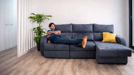 Portrait of caucasian man using smartphone lying down on couch at beautiful home interior. Caucasian man checks phone device on the sofa of house.の写真素材