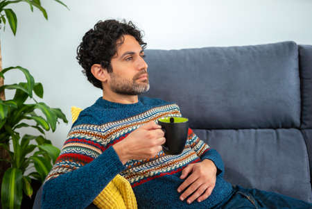 Portrait of cool and handsome man drinking morning expresso coffee and sitting on a couch at home interior. A person is resting and looking away.の写真素材