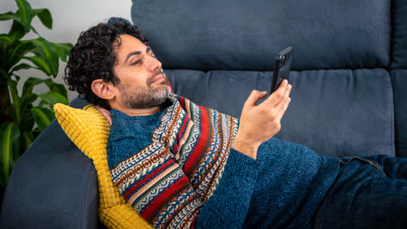 Happy handsome man smiling while using his smart phone and lying down on the couch at home. Confident bearded man relaxing and typing on smartphone at sofa of house.の写真素材