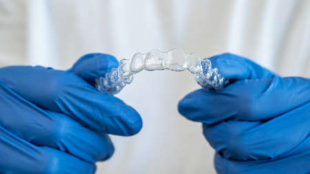 Male hand in gloves holding dental aligner retainer on white background of dental clinic for beautiful teeth treatment. Orthodontic removable straighteners in dentist officeの写真素材
