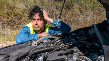 Face of unhappy male driver near a broken car with popped up hood inspecting his vehicle motor....