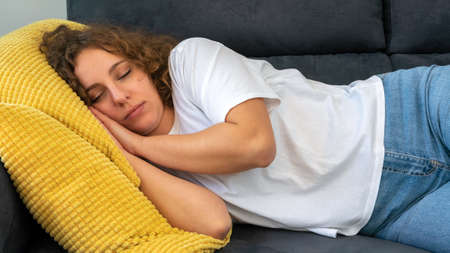 Sleeping woman with curly hair taking nap on the sofa during the day at cozy apartment. Attractive girl sleep lying on the comfortable couch with eyes closed. Dreaming and resting conceptの写真素材