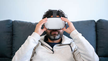 Bearded man sitting on couch and watching movie with VR glasses headset at cozy home. Play video...