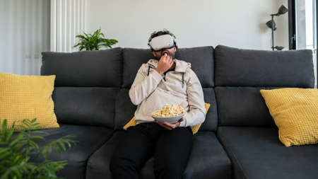 Bearded man sitting on couch and watching movie with VR glasses headset while eating popcorn at cozy home. Enjoy immersive futuristic virtual reality 3D travel tour, watch 360 videoの写真素材