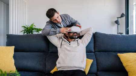 Caucasian men enjoying virtual reality glasses while sitting on sofa at cozy home. Two happy guys...