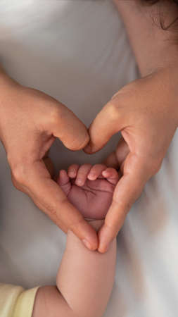 Close up mother holding hands newborn boy and giving heart signal in a room with a lot of sunlight. Baby sleeping on the mother's bed. Health care, love and relationship concept.の写真素材