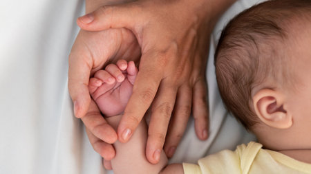 Young mother holding baby hand and showing finger and fingernail on white sheet bed. Closeup of baby sleeping on the mother's room. Family and home conceptの写真素材