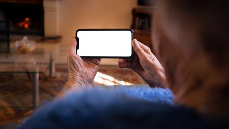 Wrinkled hands of the old woman holds a smartphone with white empty screen for your mockup. Close up...