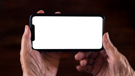 Wrinkled hands of the old woman holds a smartphone with white empty screen for your mockup. Close up...