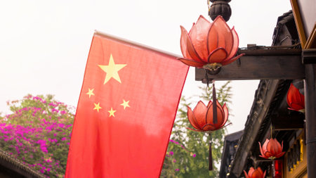 Chinese flag waves alongside red lotus shaped lanterns hanging from a traditional building. Bright pink flowers and green trees create a colorful, cultural backdropの写真素材