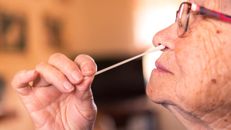 Caucasian grandma inserts a cotton swab into a nose to collect a possible positive COVID-19 sample during the pandemic. Grandmother make antigen test procedure at home.の写真素材