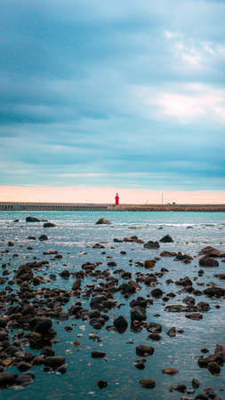 A vertical shot of a red lighthouse on a rocky beach under a cloudy skyの写真素材