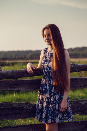 beautiful teenager girl with long healthy hair in blue dress relaxing on sunset evening in farm on summer time.の写真素材
