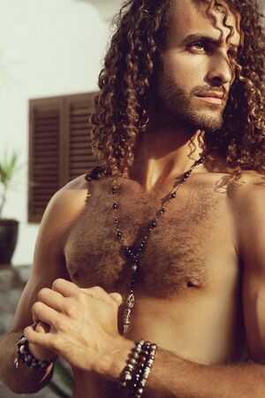 Handsome man with curly hair and jewelry on his arms and chest, outdoor photoshoot under the sun - imageの写真素材