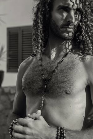 Handsome man with curly hair and jewelry on his arms and chest, outdoor photoshoot under the sun - imageの写真素材