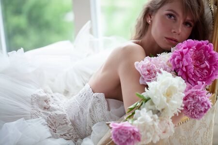 Bride in beautiful dress lying on sofa whit peonies in white studio interior. Romantic wedding style shot with sun lightの写真素材