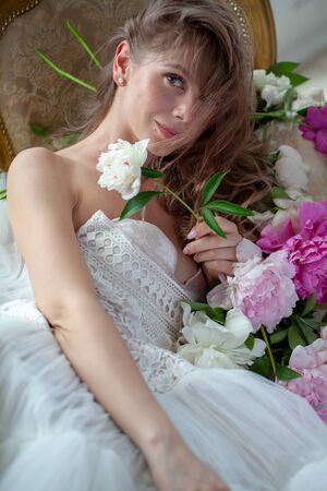 Bride in beautiful dress lying on sofa whit peonies in white studio interior. Romantic wedding style shot with sun lightの写真素材
