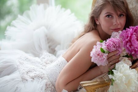 Bride in beautiful dress lying on sofa whit peonies in white studio interior. Romantic wedding style shot with sun lightの写真素材