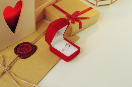 Engagement diamond ring in a red velvet box. Envelope with wax seal surrounded by gifts and a candle in the wooden box with heart shaped holeの写真素材