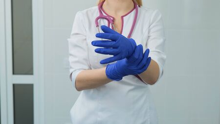 Handsome young women doctor in white medical coat, violet stethoscope putting on rubber blue gloves in hospital or clinic. Front view on a white backgroundの写真素材