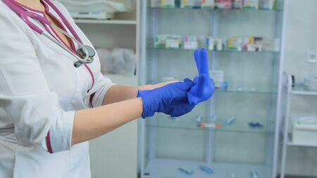 Young women Caucasian doctor in white medical coat and violet stethoscope inflates a rubber blue glove to check if it is leaky and can be used while the right glove is already wornの写真素材