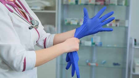 Young women doctor medic in white medical suit gown and violet stethoscope putting on rubber blue gloves on the left hand in hospital or clinic. Side viewの写真素材