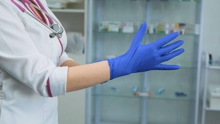 Young Caucasian female doctor in white medical coat and violet stethoscope putting on rubber blue gloves on the right hand in laboratory or hospital. Side viewの写真素材