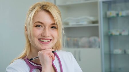 Happy young Caucasian woman blonde doctor in white medical coat and stethoscope looking at camera. Close-up face portrait of smiling female physician posing in hospital officeの写真素材