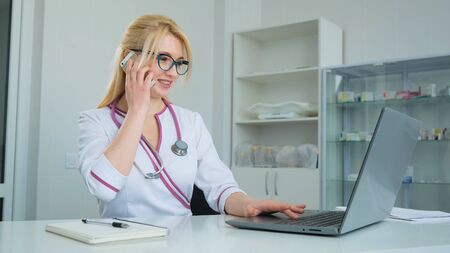 Young female Caucasian blonde doctor in lab coat and glasses using laptop and talking on mobile phone while working at the desk in medical officeの写真素材