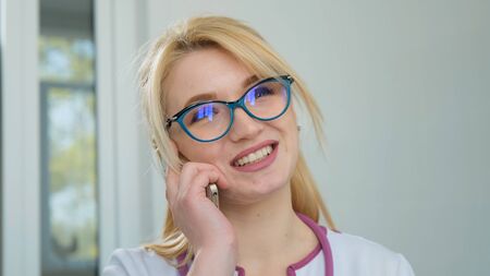 Nice blonde doctor in white coat smile and talking on phone. Positive mature woman physician talking on cell phone at clinic, window city background. To patients with care. Remote consultationの写真素材