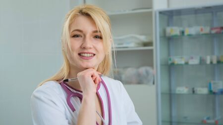 Happy young european woman blonde doctor wearing white medical coat and stethoscope raises his head and looking at camera. Smiling female physician posing in hospital officeの写真素材