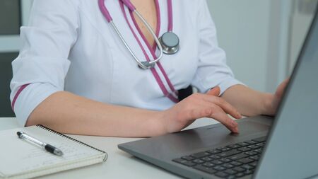 Positive Caucasian woman doctor at the hospital in white coat using laptop computer prints a recipe. Close up of Medic doing online research. Cmera movement from top to bottomの写真素材