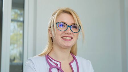 Young Caucasian female doctor in white medical coat, stethoscope and glasses looking ahead, dreaming about the future career. Close up portrait in clinicの写真素材