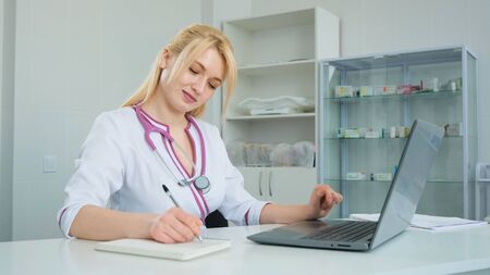 Caucasian young handsome physician sitting at table in cabinet and making notes in medical journal. The Female doctor thinks about the diagnosis and writes a prescriptionの写真素材