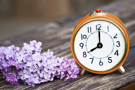 Daylight savings, spring forward concept - orange alarm clock and purple lilac flowers