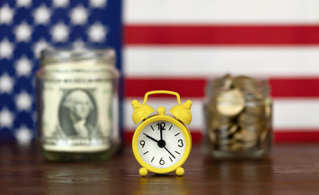 Alarm clock with US dollar bills and gold coins in jars with American flag in the background....