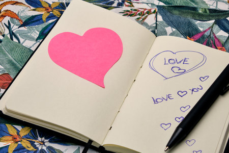 Open diary with a pen, a pink heart and written the word Love, on top of a table with a tropical...